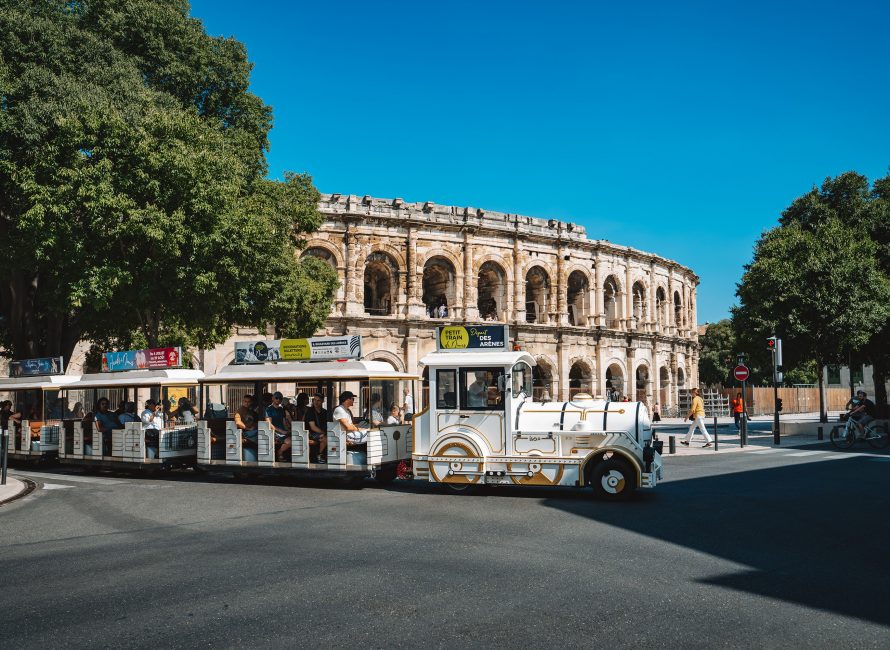 Petit train touristique circulant dans les rues de Nîmes, transportant des visiteurs à la découverte de la ville et passant devant les Arènes.