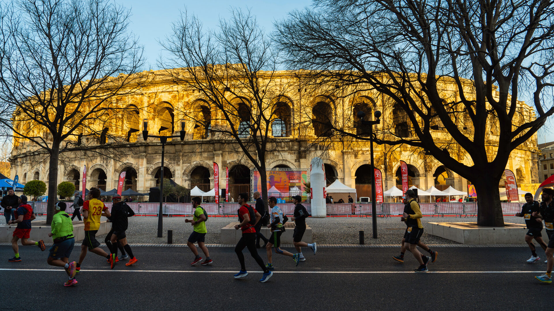 Photo de course à Nîmes, les participants passent devant les Arènes de Nîmes. Ils courent à vitesse soutenue sous un ciel bleu et un soleil descendant dans les rues de Nîmes en hiver.