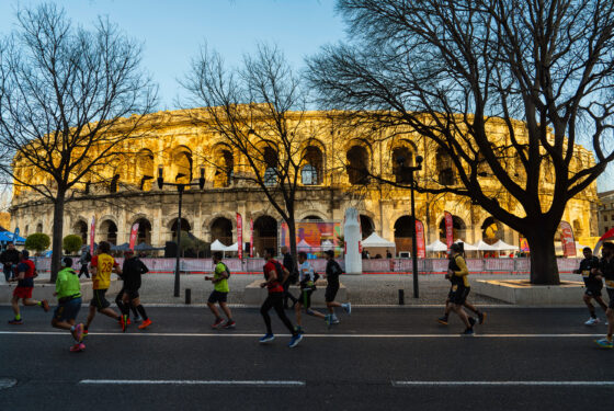 Photo de course à Nîmes, les participants passent devant les Arènes de Nîmes. Ils courent à vitesse soutenue sous un ciel bleu et un soleil descendant dans les rues de Nîmes en hiver.
