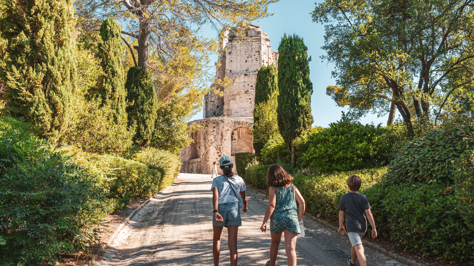 Trois enfants marchent sur un chemin bordé de verdure dans les Jardins de la Fontaine à Nîmes, en direction de la Tour Magne, sous un ciel bleu printanier.