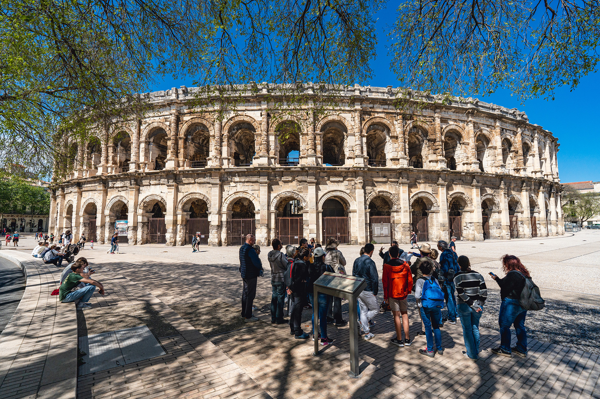Visiteurs se promenant autour des Arènes de Nîmes, amphithéâtre romain emblématique, sous un ciel ensoleillé.