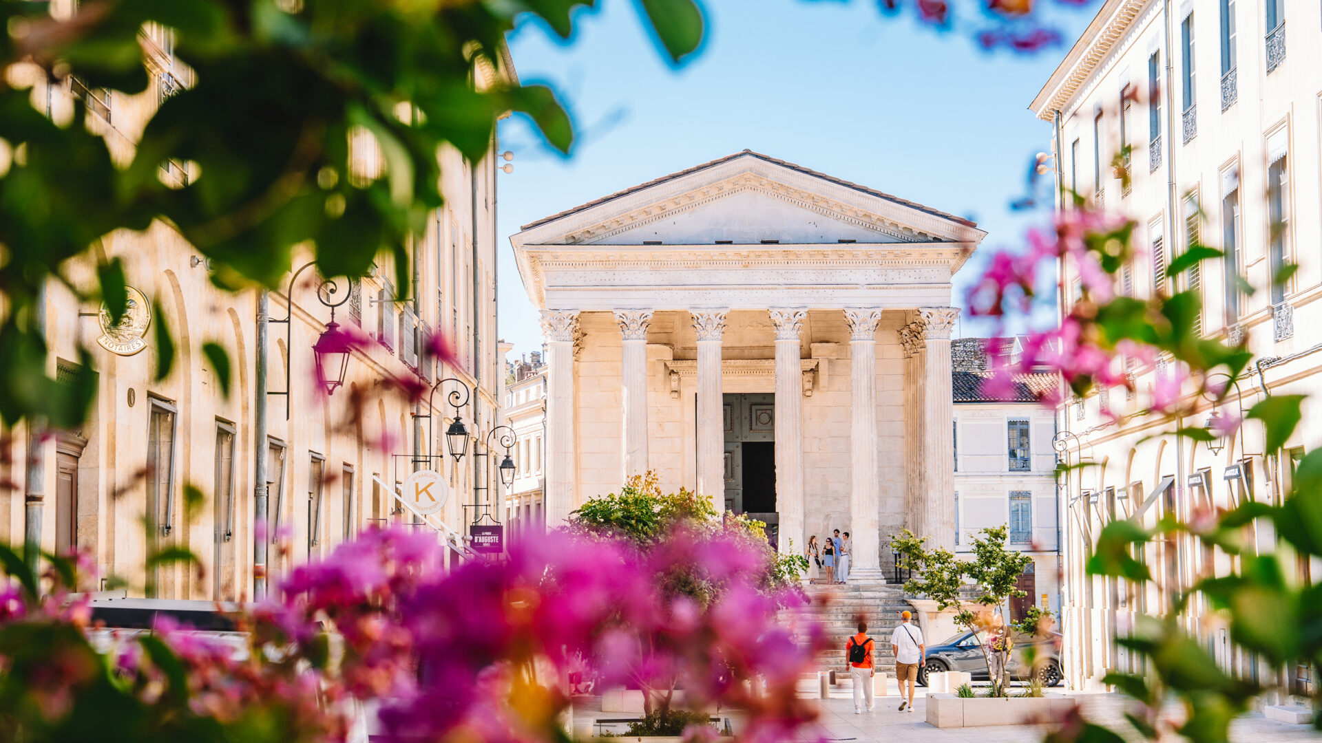 Entrée de la Maison Carrée à Nîmes, encadrée par des fleurs roses au premier plan, dans une rue ensoleillée où quelques passants se promènent sous un ciel bleu printanier.